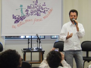 Foto de um home de barba falando ao microfone em cima de um palco.