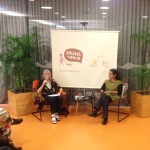 Foto com duas mulheres sentadas, uma senhora grisalha fala ao microfone e a sua esquerda uma mulher observa a senhora falando ao microfone.