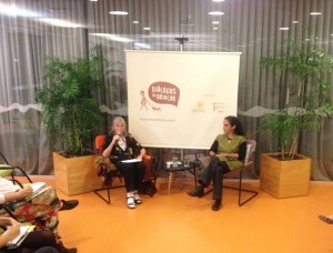 Foto com duas mulheres sentadas, uma senhora grisalha fala ao microfone e a sua esquerda uma mulher observa a senhora falando ao microfone.