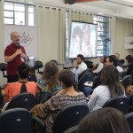 Foto composta por várias pessoas que observam um homem de barba e cabelos grisalho, esse homem está falando ao microfone.