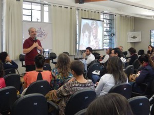 Foto composta por várias pessoas que observam um homem de barba e cabelos grisalho, esse homem está falando ao microfone.