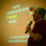 Foto ilustrando um homem falando ao microfone, com sua mão direita ele aponta para um telão, onde varias palavras se destacam, como: "Saúde, Ausência de doenças, Conexão, Olhar".