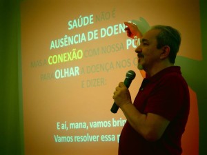 Foto ilustrando um homem falando ao microfone, com sua mão direita ele aponta para um telão, onde varias palavras se destacam, como: "Saúde, Ausência de doenças, Conexão, Olhar".