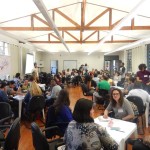 Foto de um salão, onde pessoas estão sentadas em várias mesas.