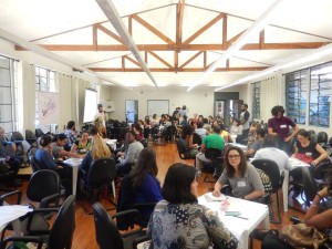 Foto de um salão, onde pessoas estão sentadas em várias mesas.