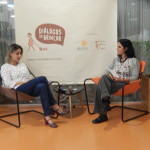 Foto de duas mulheres sentadas, a primeira mulher está olha para baixo enquanto fala, a sua direita uma mulher com folhas na mão observa a outra falar.