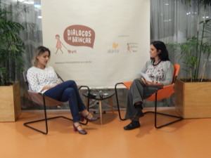 Foto de duas mulheres sentadas, a primeira mulher está olha para baixo enquanto fala, a sua direita uma mulher com folhas na mão observa a outra falar.