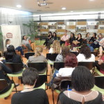 Foto composta por várias pessoas sentadas em círculo, ao fundo duas mulheres estão sentadas, ambas de pernas cruzadas, a primeira está falando com as pessoas, enquanto a sua esquerda a outra mulher segura o microfone com sua mão direita enquanto com a esquerda mexe em seu cabelo.