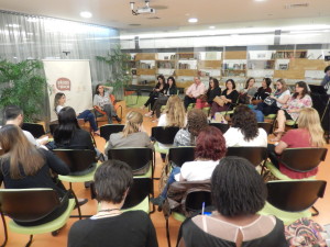 Foto composta por várias pessoas sentadas em círculo, ao fundo duas mulheres estão sentadas, ambas de pernas cruzadas, a primeira está falando com as pessoas, enquanto a sua esquerda a outra mulher segura o microfone com sua mão direita enquanto com a esquerda mexe em seu cabelo.