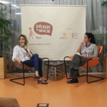 Foto composta por duas mulheres, de pernas cruzadas fala ao microfone, em seu lado esquerdo outra mulher a observar falar.