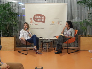 Foto composta por duas mulheres, de pernas cruzadas fala ao microfone, em seu lado esquerdo outra mulher a observar falar.