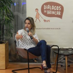 Foto de uma mulher sentada, com suas pernas cruzadas, segura um microfone com sua mão esquerda, enquanto fala ao microfone, gesticula com sua mão direita.