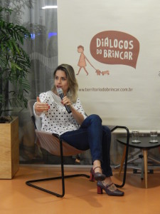 Foto de uma mulher sentada, com suas pernas cruzadas, segura um microfone com sua mão esquerda, enquanto fala ao microfone, gesticula com sua mão direita.