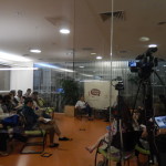 Foto composta por várias pessoas sentadas observando uma mulher falando ao microfone, uma câmera está filmando.