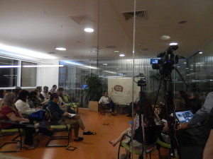 Foto composta por várias pessoas sentadas observando uma mulher falando ao microfone, uma câmera está filmando.