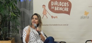 Foto de uma mulher sentada, com suas pernas cruzadas, fala ao microfone.