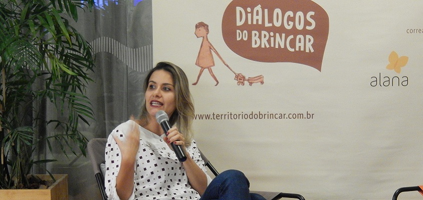 Foto de uma mulher sentada, com suas pernas cruzadas, fala ao microfone.