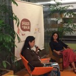 Foto composta por duas mulheres, uma fala ao microfone olhando para baixo, enquanto a sua esquerda outra mulher a observa.