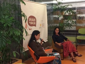 Foto composta por duas mulheres, uma fala ao microfone olhando para baixo, enquanto a sua esquerda outra mulher a observa.