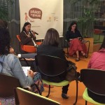 Foto composta por várias pessoas sentadas, ao fundo duas mulheres sentadas, uma fala ao microfone enquanto a sua direita a outra mulher esta lendo um caderno em sua mão, a plateia observam as duas mulheres.