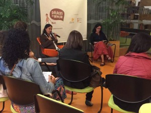 Foto composta por várias pessoas sentadas, ao fundo duas mulheres sentadas, uma fala ao microfone enquanto a sua direita a outra mulher esta lendo um caderno em sua mão, a plateia observam as duas mulheres.