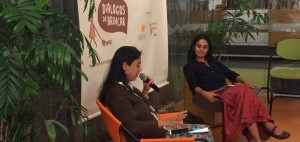 Foto composta por duas mulheres, uma fala ao microfone olhando para baixo, enquanto a sua esquerda outra mulher a observa.