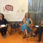 Foto composta por três mulheres sentadas, a mulher da esquerda veste preto e está segundo folhas em suas mãos, a sua esquerda uma mulher veste um vestido azul e está de pernas cruzadas, a esquerda da mulher de azul tem uma mulher vestida de multicolorido de pernas cruzadas e segundado folhas em suas mãos, ambas as três estão sorrindo.