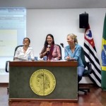 Foto composta por três mulheres sentadas em uma mesa em cima de um palco, a mulher da esquerda veste branco, a mulher que está no meio veste rosa e fala ao microfone, a mulher que está à direita veste azul e observa a mulher de rosa, as três estão sorrindo, ao fundo delas tem três bandeiras e um telão com informações.