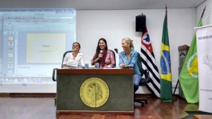 Foto composta por três mulheres sentadas em uma mesa em cima de um palco, a mulher da esquerda veste branco, a mulher que está no meio veste rosa e fala ao microfone, a mulher que está à direita veste azul e observa a mulher de rosa, as três estão sorrindo, ao fundo delas tem três bandeiras e um telão com informações.