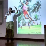 Foto de uma mulher em um palco falando ao microfone que segura com a sua mão direita e gesticulando com a sua mão esquerda, de costa para uma foto retroprojetora de crianças brincando.