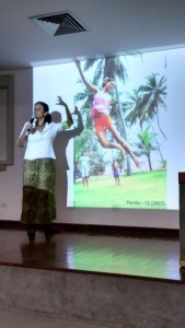 Foto de uma mulher em um palco falando ao microfone que segura com a sua mão direita e gesticulando com a sua mão esquerda, de costa para uma foto retroprojetora de crianças brincando.
