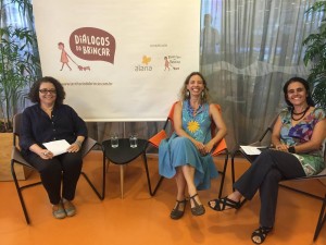 Foto composta por três mulheres sentadas, a esquerda da foto uma mulher veste preto e segura folhas em sua mão, entre a mulher de preto e mulher que está no meio tem uma mesa com dois copos de agua, a mulher do meio veste azul e a direita da foto temos uma mulher que veste multicolorido, as três estão sorrindo.