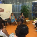 Foto de três mulheres dando palestra em uma roda de pessoas a mulher ao fundo da foto fala ao microfone enquanto a sua direita uma mulher vestida de azul a observa.