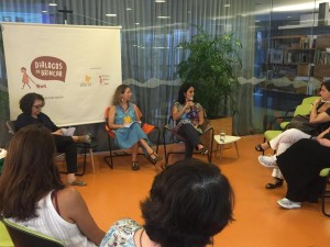 Foto de três mulheres dando palestra em uma roda de pessoas a mulher ao fundo da foto fala ao microfone enquanto a sua direita uma mulher vestida de azul a observa.