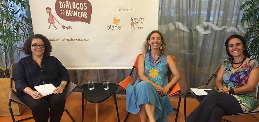 Foto composta por três mulheres sentadas, a esquerda da foto uma mulher veste preto e segura folhas em sua mão, entre a mulher de preto e mulher que está no meio tem uma mesa com dois copos de agua, a mulher do meio veste azul e a direita da foto temos uma mulher que veste multicolorido, as três estão sorrindo.