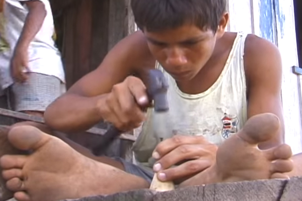 Foto de um menino descalço martelando com um prego a base de um peão de brinquedo.