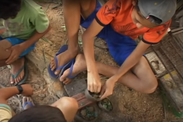 Foto de quatro crianças sentadas em círculo em um chão de terra, o primeiro garoto veste uma camisa laranja e um boné cinza está separando pedras dentro de uma tigela em cima de uma pedra, a sua esquerda um garoto está de lado para a roda o último observa o garoto de camisa laranja.