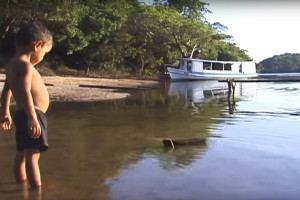 Foto de um garoto dentro de um ribeirão olha para um toco de madeira que boia a sua frente.
