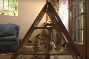 Uma menina brinca de casinha com uma casa de brinquedo feita de madeira, na casinha tem vários objetos em suas salas.
