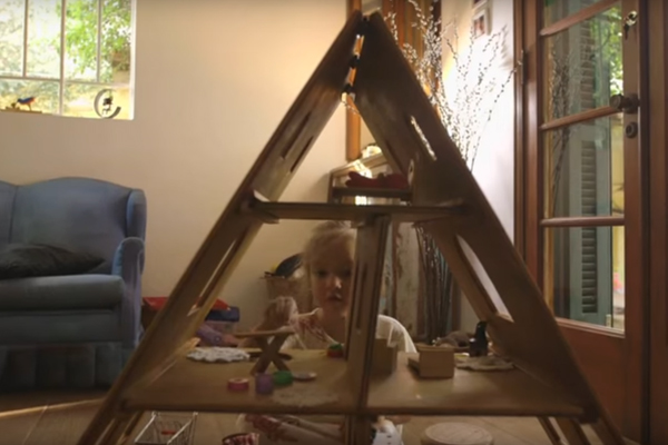 Uma menina brinca de casinha com uma casa de brinquedo feita de madeira, na casinha tem vários objetos em suas salas.