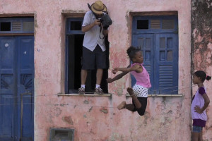 Foto composta por três pessoas, duas meninas negras brincando em frente de uma casa, enquanto uma salta a outra observa, e um homem de chapéu bege tira uma foto em cima de uma das duas janelas.