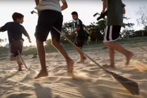 Quatro garotos correm em um chão de areia, em cima de cabos de gralho de uma arvore brincão de montar em cavalo.