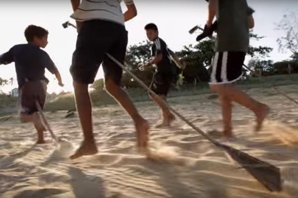 Quatro garotos correm em um chão de areia, em cima de cabos de gralho de uma arvore brincão de montar em cavalo.