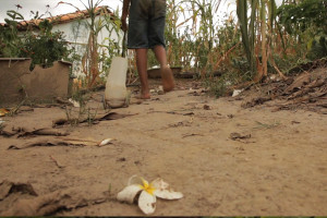 Foto de um garoto andando em um chão de terra, ele está de costas caminhando para longe da foto, uma casca de banana está jogada ao chão, o garoto puxa um carrinho de mão com sua mão esquerda, somente é possível ver a cintura para baixo do garoto.