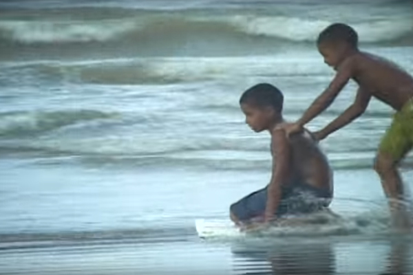 Foto de dois garotos negos, ambos estão sem camisa e brincando na beira do mar, em pé um garoto com bermudas verde, correndo empurra o outro garoto de bermuda azul, o garoto de bermuda azul está ajoelhado em uma prancha.