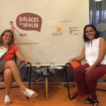 Foto composta por duas mulheres sentadas e sorrindo, a mulher da esquerda cruza as pernas.