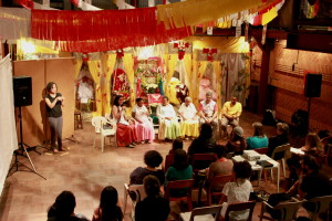 Foto ilustrando  o público total de uma palestra ,onde ao fundo há um palco composto de quatro mulheres e dois homens falando a um número de pessoas sentadas assistindo ao evento.