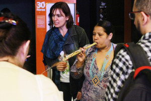 Foto ilustrando interação entre pessoas , onde ao centro á duas mulheres onde uma delas está tocando uma flauta indígena.