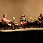 Foto ilustrando uma palestra , onde em um palco há uma mulher ao centro com um microfone á mão , á esquerda á um homem e a direita á outra mulher.