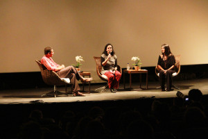 Foto ilustrando uma palestra , onde em um palco há uma mulher ao centro com um microfone á mão  , á esquerda á um homem e a direita á outra mulher.